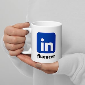 May include: White ceramic mug featuring the LinkedIn logo in a blue square with the word "influencer" in black text below. The mug has a curved handle and is held by a person.