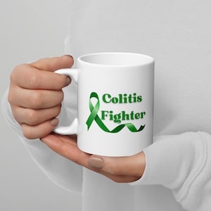 May include: White ceramic mug with a green ribbon and the words "Colitis Fighter" printed on the side. The mug has a curved handle and is held by a person.
