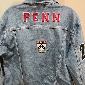 May include: A blue denim jacket with a pink "PENN" patch on the back, a crest with the words "Sine Moribus" and a black number "2" on the sleeve.