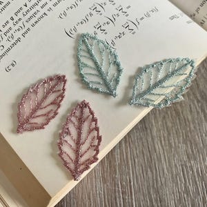May include: Three delicate lace leaf embellishments in pink and blue. The leaves are on a book with mathematical equations.