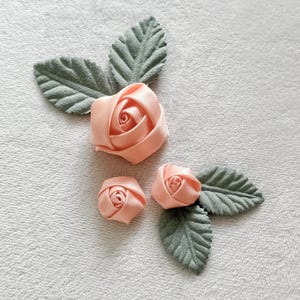 Op de afbeelding: Perzikroze satijnen rozen met groene stoffen bladeren gerangschikt op een wit, getextureerd oppervlak. De opstelling omvat een grote roos en twee kleinere rozenknoppen, met drie bladeren. De rozen zijn gemaakt van gevouwen satijn.