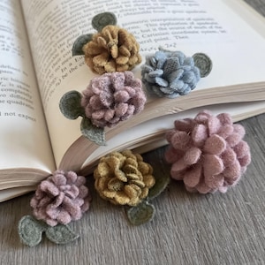 May include: Five felt flower embellishments in shades of pink, blue, and yellow. The flowers have a textured, fluffy appearance and are arranged on a wooden surface.
