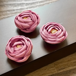 Op de afbeelding: Drie handgemaakte, stoffen bloemversieringen in oudroze met een gelaagd bloemblaadjesontwerp en een klein, goudkleurig centrum. De bloemen zijn gerangschikt op een donkergrijze ondergrond, met een houten achtergrond.