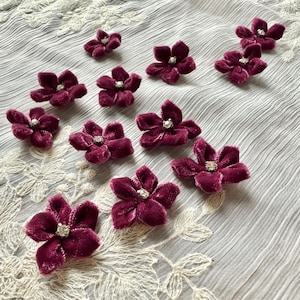 May include: Deep burgundy velvet flower appliques with a central rhinestone. The flowers are arranged on a textured, light grey fabric with white lace trim. These floral embellishments are suitable for craft projects and fabric decoration.