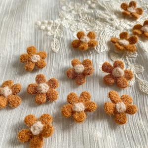 May include: A collection of ten felt flower embellishments in shades of brown and cream. The flowers have a simple, five-petal design and a small, white center.