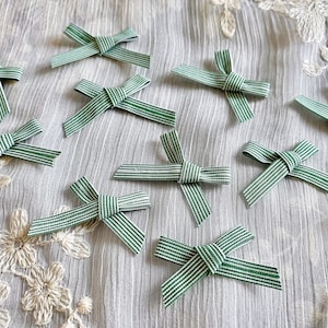May include: A collection of 10 green and white striped fabric bows. The bows are arranged on a white fabric background.