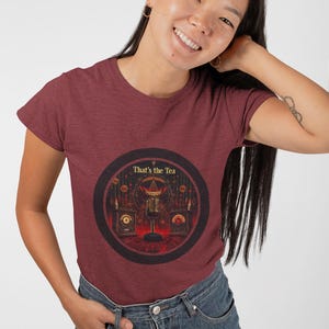 Op de afbeelding: Een maroon t-shirt met een afbeelding van een microfoon in een donkere kamer met kaarsen en de tekst "That's the Tea".