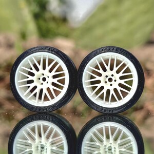 May include: Four miniature car wheels with white spokes and black tires are displayed. The tires have the text "PROXES" and "TOYO" printed on them. The wheels are reflected in the surface below.