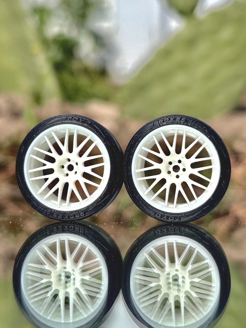 May include: Four miniature car wheels with white spokes and black tires are displayed. The tires have the text "PROXES" and "TOYO" printed on them. The wheels are reflected in the surface below.