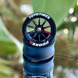 May include: A black toy car wheel with the text "TOYO TIRES" in white. The wheel has red accents and is stacked on three black tyres with "PROXES" on the sidewall. The background is out of focus.