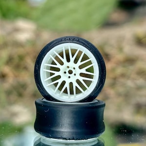 May include: Two model car tires stacked on top of each other. The top tire has a white spoke design and the word "TOYO" printed on the tire. The bottom tire is black.