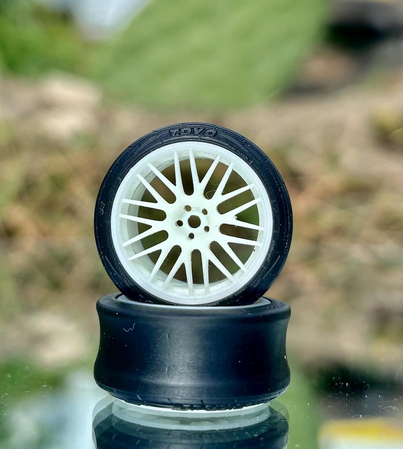 May include: Two model car tires stacked on top of each other. The top tire has a white spoke design and the word "TOYO" printed on the tire. The bottom tire is black.