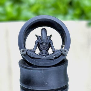 Puede incluir: Tapon de oreja negro con un diseño de alienígena meditando. El alienígena está sentado en posición de loto dentro de un marco circular. El tapón es de material oscuro, para piercings estirados.