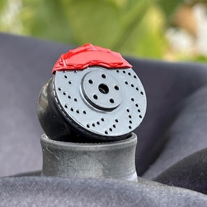 May include: A close-up of a black brake rotor with a red brake caliper. The rotor has a series of holes drilled into it for ventilation. The caliper is mounted on top of the rotor.