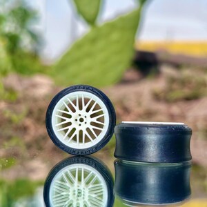 May include: Two miniature black and white model car tires with a white spoke wheel design. The tires are set against a reflective surface, with a blurred green background.