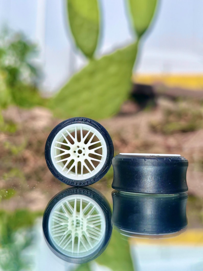 May include: Two miniature black and white model car tires with a white spoke wheel design. The tires are set against a reflective surface, with a blurred green background.