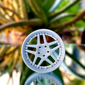 May include: A gray plastic model car wheel with a multi-spoke design. The wheel has a reflective surface and is sitting on a glass surface.