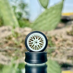 May include: A miniature black and white car wheel with a white spoke design is stacked on top of a black cylinder. The wheel is set against a blurred background of green foliage and a light blue sky.