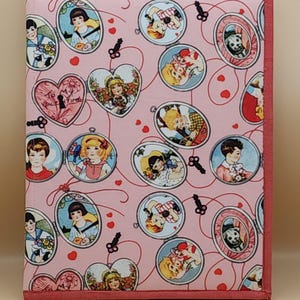May include: A pink fabric journal cover with a repeating pattern of vintage-style portraits in oval frames and heart-shaped lockets. The cover is trimmed with a darker pink border and features a stitched binding.