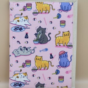 May include: A pink fabric notebook cover featuring a repeating pattern of cartoon cats, sushi, and bowls of noodles. The cats are in various colors and poses. The cover has a cream-colored border.