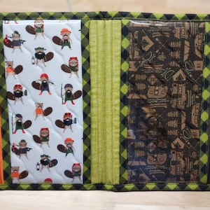 May include: A green and black checkered fabric wallet with a zipper closure and a clear plastic window. The front of the wallet features a repeating pattern of beavers wearing hats and holding axes. The back of the wallet features a repeating pattern of camping-related icons.