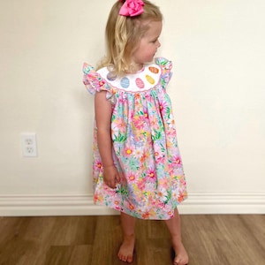 May include: A little girl wearing a white dress with a pink and green floral print and a white collar with colorful Easter eggs. She is wearing a pink bow in her hair.