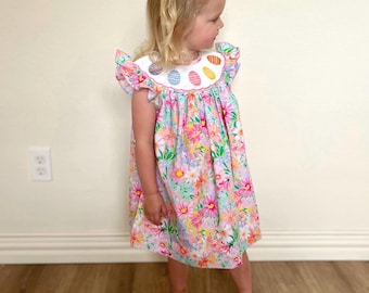 May include: A little girl wearing a white dress with a pink and green floral print and a white collar with colorful Easter eggs. She is wearing a pink bow in her hair.