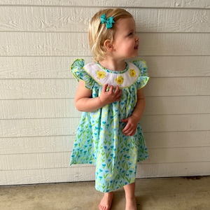 May include: A little girl wearing a light blue dress with a white collar and yellow chicks embroidered on the collar. The dress has a floral pattern with blue and yellow flowers. The girl has blonde hair and is wearing a turquoise bow in her hair.