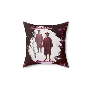 May include: Square pillow with a dark burgundy background and a white brushstroke design. The pillow features two graduates in maroon graduation gowns and the text "Congratulations", "Class of 2026", "Put Name Here", and "Graduate".