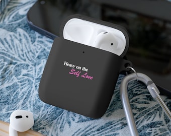 May include: A matte black silicone protective case for wireless earbuds featuring the white and pink text "Heavy on the Self Love" printed on the front. The case is open, revealing white earbuds inside, and includes a silver metal carabiner clip.