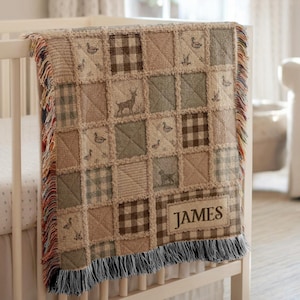 Puede incluir: Una manta de patchwork tejida sobre una cuna blanca, con una cuadrícula de cuadrados en tonos beige, marrón y verde salvia. El diseño incluye motivos de ciervos, patos y cuadros, con el nombre JAMES bordado en un parche rectangular.