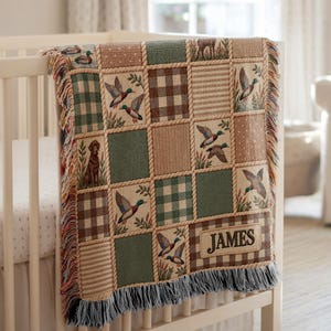 May include: A woven throw blanket featuring a patchwork design of mallard ducks, a dog, and brown and green plaid patterns. The blanket includes a rectangular patch with the name JAMES embroidered in dark brown thread and has fringed edges.