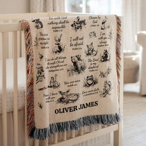 May include: A beige baby blanket with black illustrations of animals and Bible verses. The blanket is draped over a white crib. The name "OLIVER JAMES" is printed on the blanket.