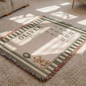 May include: A rectangular woven blanket featuring a cream-coloured centre with the name Oliver James printed in dark green. The design includes a brown teddy bear holding a green balloon and a list of numbers 1 through 12 labelled as MONTHS.