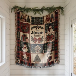 May include: A woven throw blanket featuring a grid of Christmas-themed illustrations including a reindeer, teddy bear, snowman, house, gifts, and a star. The centre panel displays the text The Johnson's Family surrounded by pine branches and a red bow.