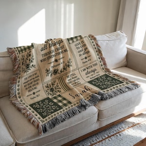May include: A woven throw blanket featuring a cream and dark green color palette with a checkered border and fringe edges. The blanket displays floral illustrations and religious text including Philippians 4:13, Psalm 46:10, and the name Sarah.