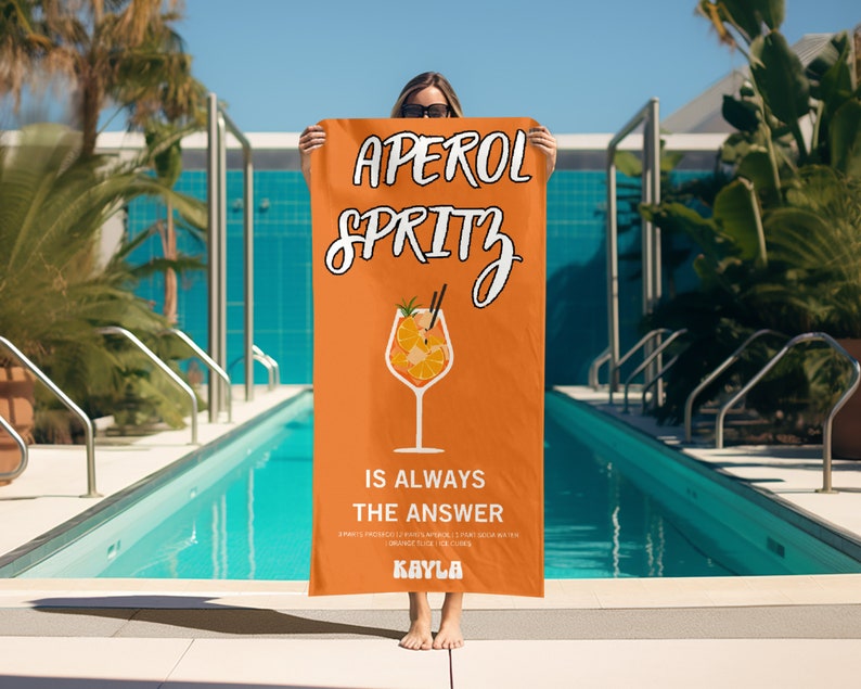 May include: Orange and white beach towel with the text "Aperol Spritz is always the answer" and a graphic of an Aperol Spritz cocktail. The towel is held by a person in front of a swimming pool.