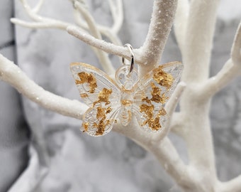 Puede incluir: Un amuleto de mariposa de resina transparente con detalles de pan de oro. El amuleto cuelga de un lazo plateado.