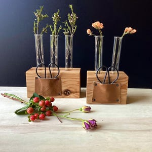 May include: Two wooden block vases with glass tubes holding flowers. Each block has a leather pocket and a pair of black scissors. Berries and a purple rose are in the foreground. The background is dark.