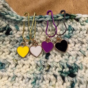 May include: Four heart-shaped stitch markers in yellow, white, purple, and black, each attached to a safety pin. The markers are hanging from a crocheted piece of fabric in shades of white, green, and blue.