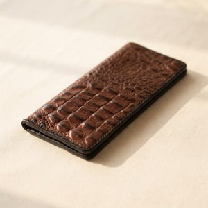 Può includere: Un portafoglio rettangolare e sottile con un esterno marrone scuro caratterizzato da una texture in rilievo che imita la pelle di coccodrillo. Il portafoglio è ripiegato, mostrando un design a scaglie uniforme e bordi scuri e ben definiti.