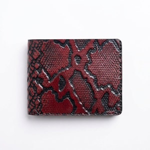 Puede incluir: Una cartera rectangular plegable con un patrón texturizado de piel de serpiente en rojo oscuro y negro. La superficie muestra un diseño repetitivo de escamas con contornos negros que separan los parches de color rojo vibrante.