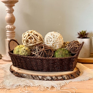 May include: A brown woven basket filled with decorative spheres of various textures and colors. The basket sits on a wooden slice atop a burlap cloth. The spheres include woven, moss-covered, and twig-like designs, creating a natural, rustic aesthetic.
