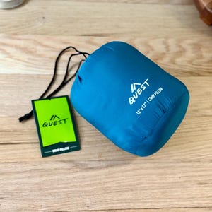 Puede incluir: Una almohada de viaje azul turquesa con cierre de cordón. La almohada tiene la palabra "QUEST" y un logotipo de montaña impreso. La almohada mide 18" x 12" y está etiquetada como "Camp Pillow".