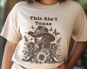 Puede incluir: Una camiseta beige con un estampado gráfico de una mujer con un sombrero de vaquero y botas. El texto "This Ain't Texas" está impreso en la camiseta.