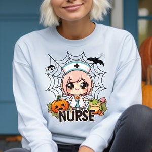 Puede incluir: Una sudadera azul claro con un personaje de dibujos animados de una enfermera que lleva un vestido blanco y un gorro rosa. El personaje está rodeado de una telaraña con una araña, un murciélago, una calabaza y una rana. El texto "NURSE" está impreso debajo del personaje.