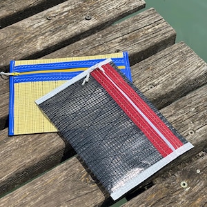 May include: Two zippered pouches rest on a weathered wooden dock. One is yellow with a blue border and zipper, the other is black with a red and white stripe. Both have a textured, grid-like pattern.