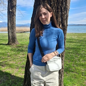 May include: A person wearing a blue turtleneck sweater and cream-colored corduroy pants. A white crossbody bag with a black strap is worn over the shoulder. The background includes a tree, grass, and a body of water.