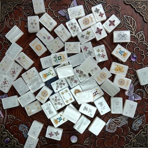 K&ouml;nnte beinhalten: Eine Sammlung wei&szlig;er, rechteckiger Mahjong-Spielsteine mit verschiedenen bunten Illustrationen, darunter Blumenmuster, Kronen, das Wort Joker und chinesische Schriftzeichen, verstreut auf einer dunkelbraunen Holzoberfl&auml;che mit floralen Schnitzereien.