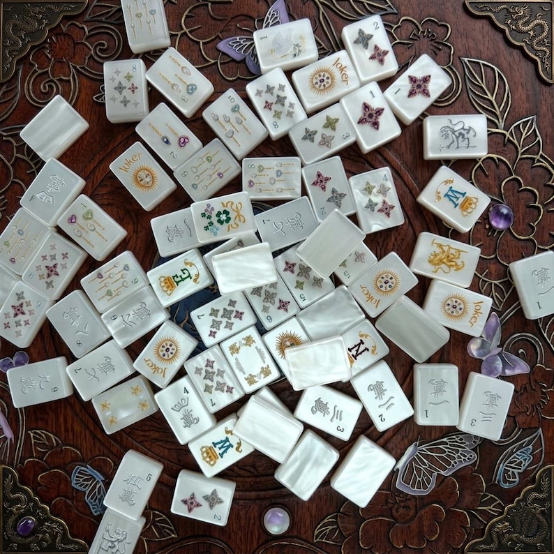 K&ouml;nnte beinhalten: Eine Sammlung wei&szlig;er, rechteckiger Mahjong-Spielsteine mit verschiedenen bunten Illustrationen, darunter Blumenmuster, Kronen, das Wort Joker und chinesische Schriftzeichen, verstreut auf einer dunkelbraunen Holzoberfl&auml;che mit floralen Schnitzereien.